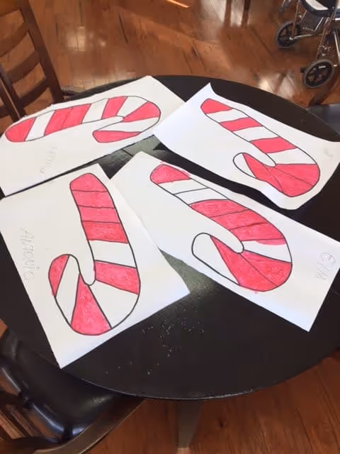 A round black table with four sheets of paper on it, each featuring a hand-drawn and colored candy cane with red and white stripes. The names 'Antonio' and 'Ethan' are visible on two of the papers. The setting appears to be indoors with wooden flooring and a wheelchair partially visible in the background.