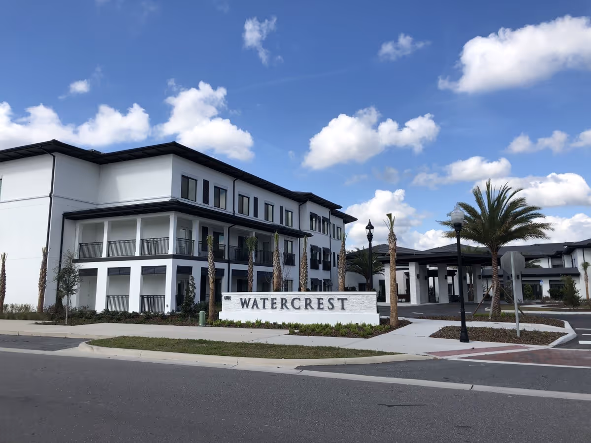 Exterior view of a modern three-story building with white walls and black trim under a blue sky with scattered clouds. The building has balconies and is surrounded by palm trees and landscaped greenery. A white sign in front reads 'WATERCREST'.