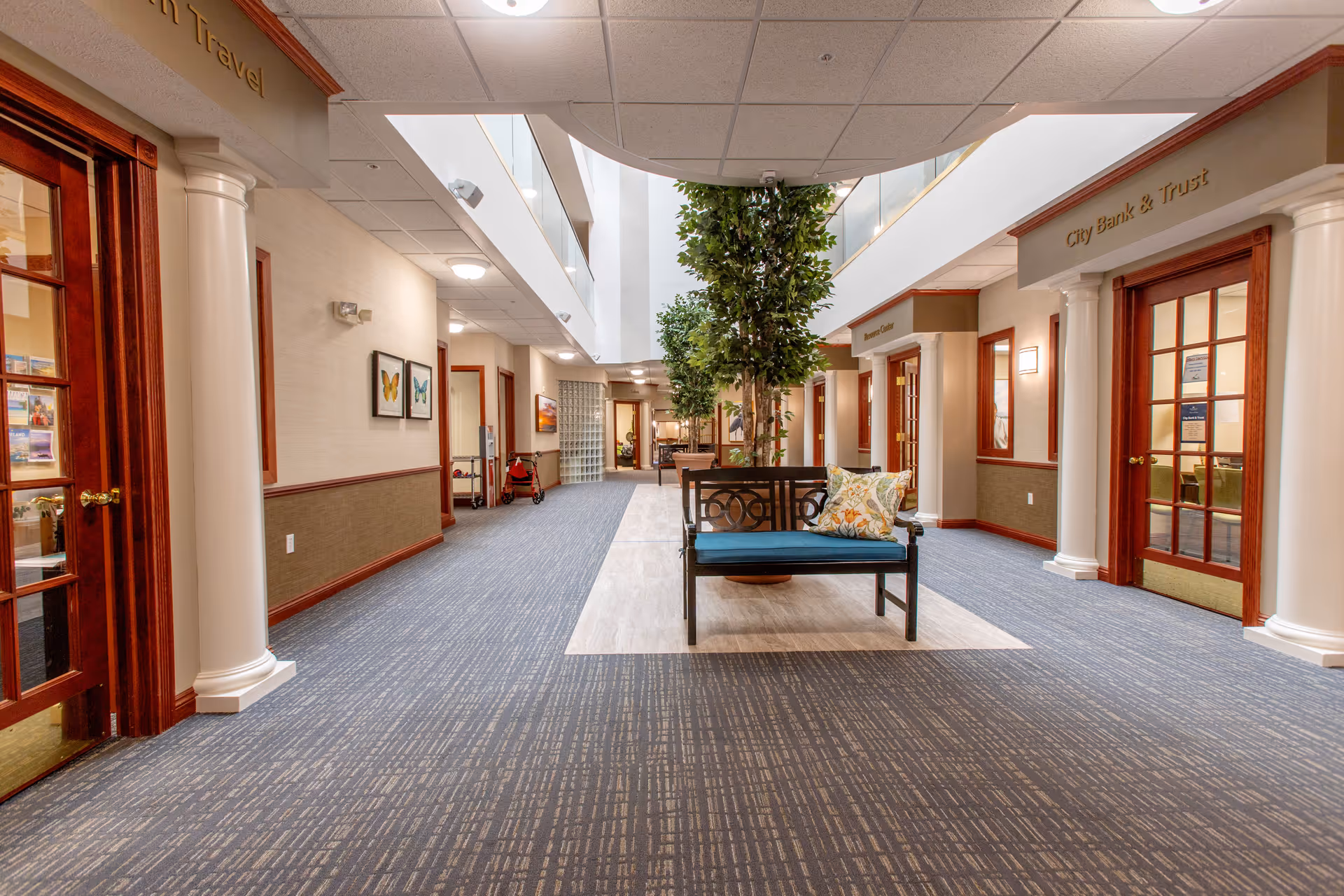 Spacious atrium-style interior corridor of a retirement community with a central bench, potted tree, and doorways to offices on either side.