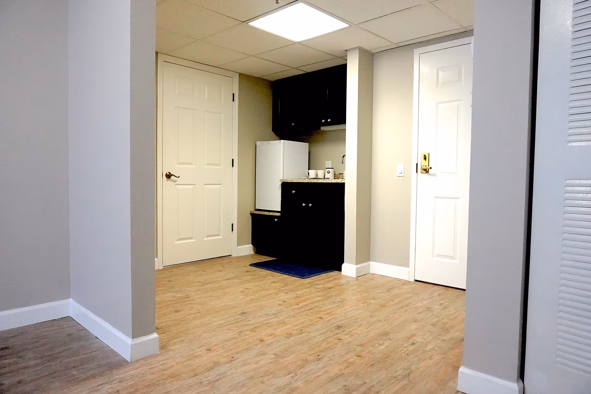 Small interior with a kitchenette featuring a mini refrigerator, sink and cabinets between two doors on wood flooring.