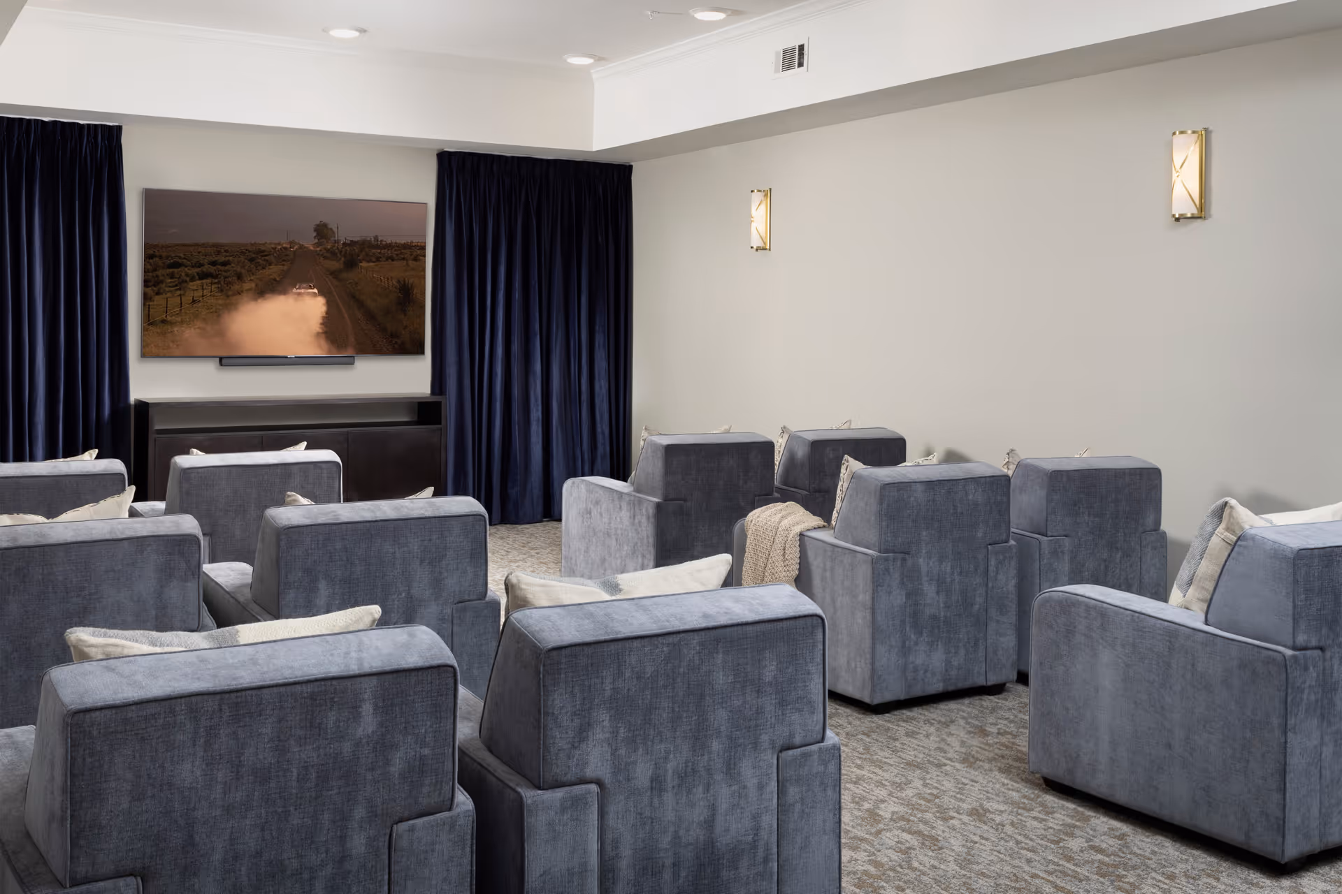 A small theater room with multiple rows of gray upholstered armchairs facing a large flat-screen TV mounted on the wall. The room has dark curtains on either side of the TV, beige walls with two wall sconces, and a carpeted floor.