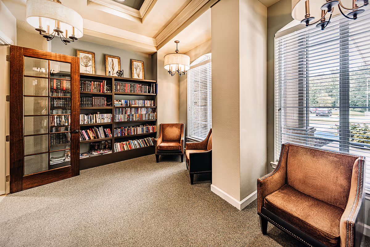 A cozy reading nook with a large bookshelf filled with books, three brown leather armchairs, and large windows with blinds letting in natural light. The room features a wooden door with glass panels and two ceiling light fixtures with fabric shades.
