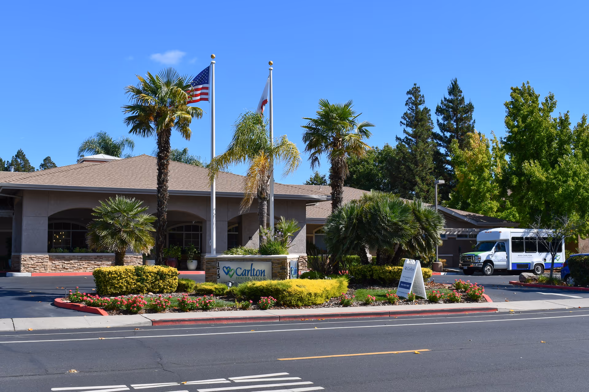 Exterior view of Carlton Senior Living Downtown Pleasant Hill facility with a single-story building, palm trees, and landscaping including bushes and flowers. Two flagpoles display the American flag and the California state flag. A white shuttle bus is parked on the right side near the building.
