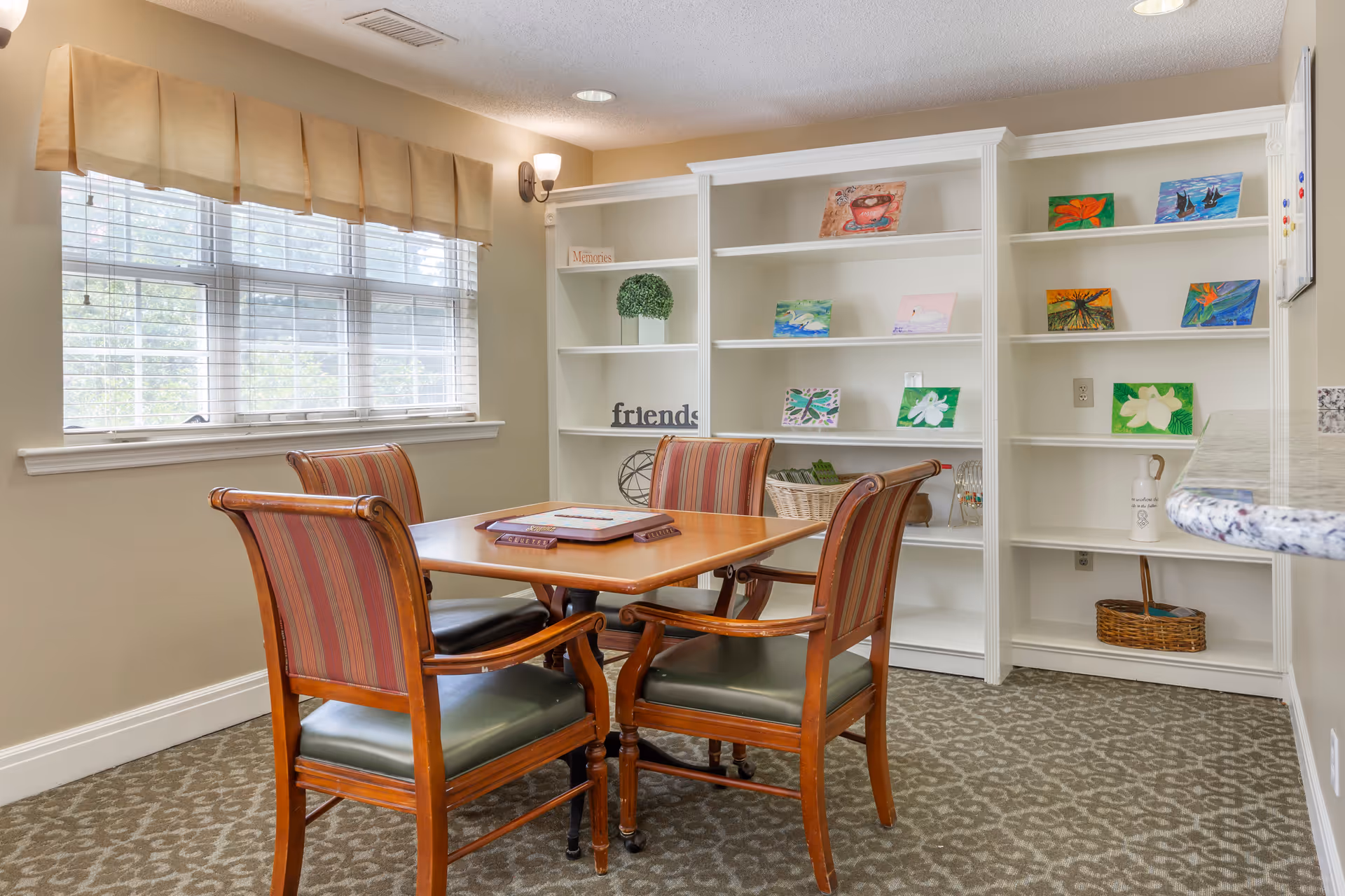 A cozy room with a wooden table and four striped upholstered chairs. Behind the table is a white built-in bookshelf displaying small colorful paintings, decorative items, and a basket. A window with beige valance and blinds lets in natural light, and the room has beige walls and patterned carpet.