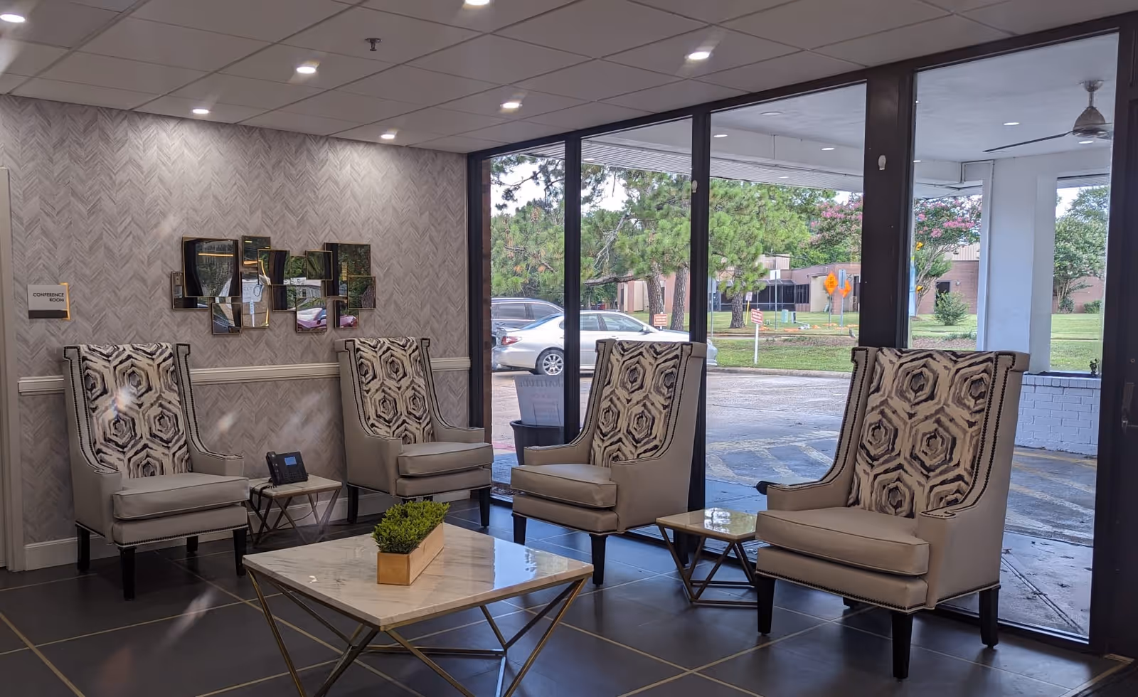 Lobby seating area with four patterned high-back chairs, small side tables and a marble-top coffee table in front of large windows overlooking the parking area.