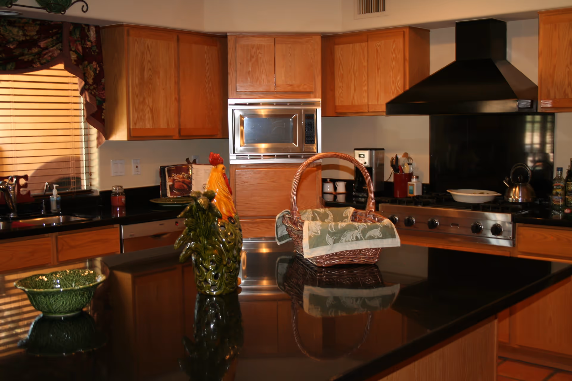 A kitchen with wooden cabinets, a black countertop island with a decorative ceramic rooster and a wicker basket with a cloth inside. The kitchen features a stainless steel microwave, a stove with a black range hood, a coffee maker, and various kitchen utensils. A window with wooden blinds and floral valance is visible on the left side.
