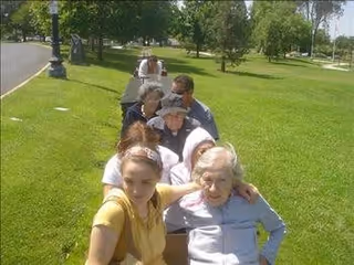 A group of elderly people and a younger woman sitting closely together outdoors on a sunny day, with green grass and trees in the background.