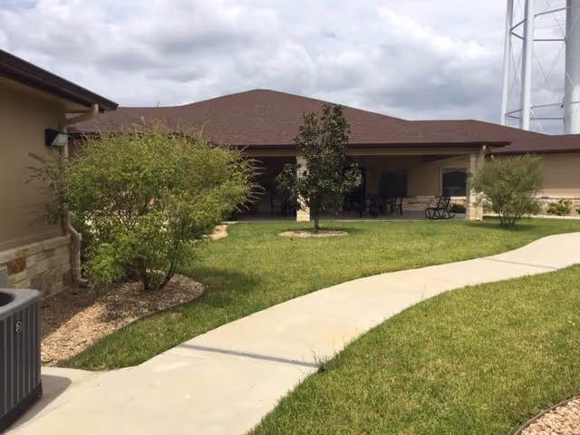 Outdoor view of a single-story building with a brown roof and beige walls, surrounded by green grass, small trees, and bushes. A curved concrete walkway leads to a covered patio area with tables and chairs. A water tower is visible in the background.