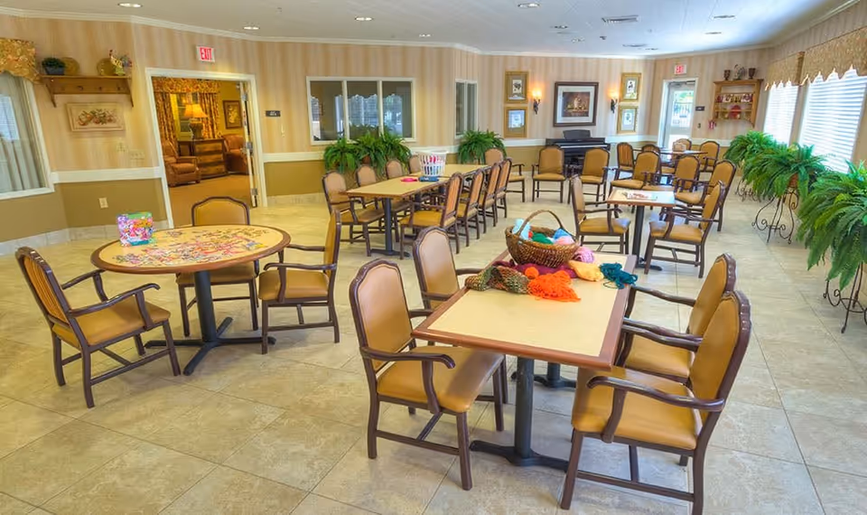A spacious, well-lit common room with multiple tables and chairs arranged for group activities. One table has a puzzle in progress, and another has a basket with colorful yarn and knitting supplies. The room features large windows with blinds, potted plants, framed artwork on the walls, and a piano in the background.