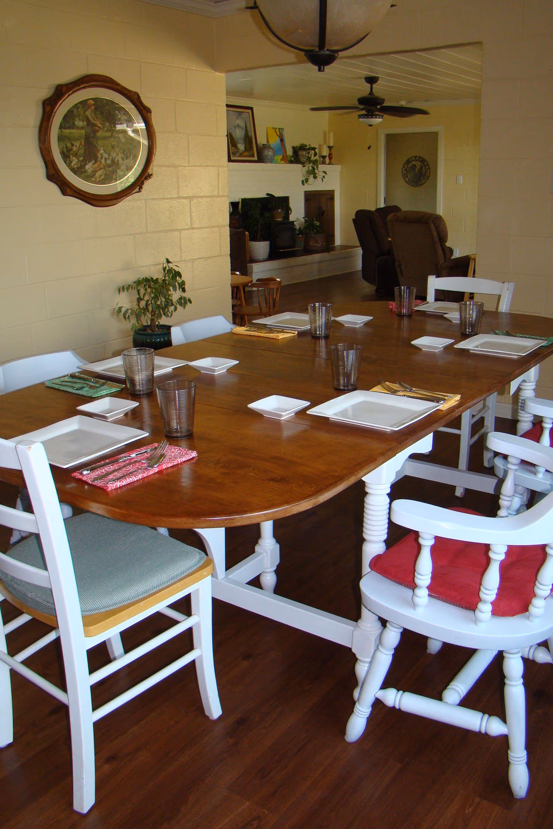 A wooden dining table set with square white plates, small square dishes, and clear drinking glasses. The table is surrounded by white chairs with cushions in various colors. In the background, there is a living area with brown recliners, a ceiling fan, and a fireplace with decorative items on the mantel. A round framed picture hangs on the beige wall to the left.