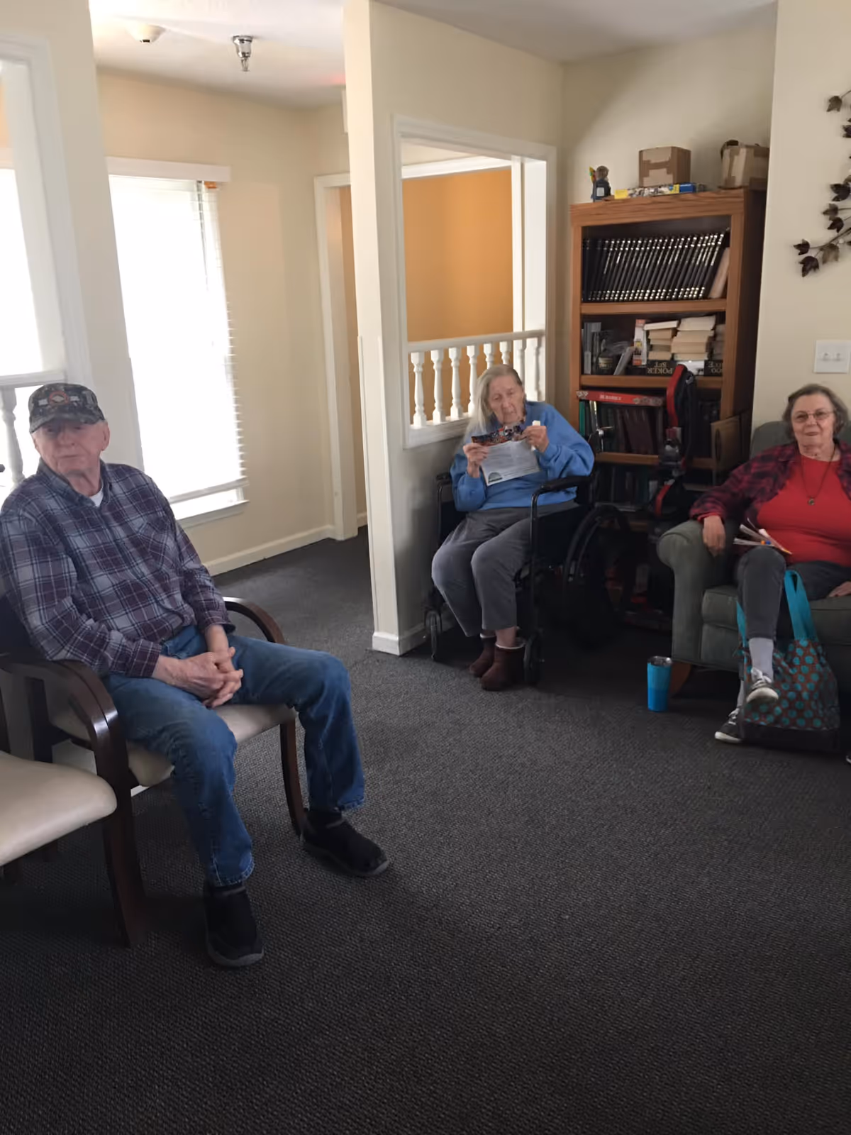 Three elderly individuals sitting in a room with carpeted floor and light-colored walls. One man is seated on a chair wearing a plaid shirt and cap, an elderly woman in a wheelchair is reading a paper, and another woman is sitting on a couch holding a book. There is a wooden bookshelf filled with books and some decorative items in the background.