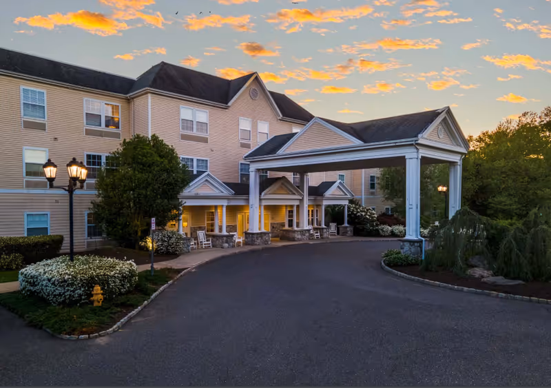 Exterior view of Monarch Spring Meadows facility at sunset, showing a large beige building with multiple windows, a covered entrance with white columns, landscaped bushes, a fire hydrant, and street lamps.