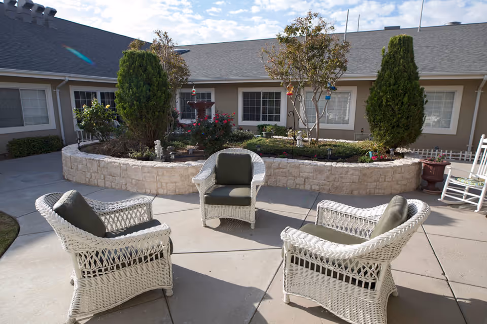 Outdoor courtyard with four white wicker chairs arranged around a paved area facing a raised stone planter bed in front of a single-story building.
