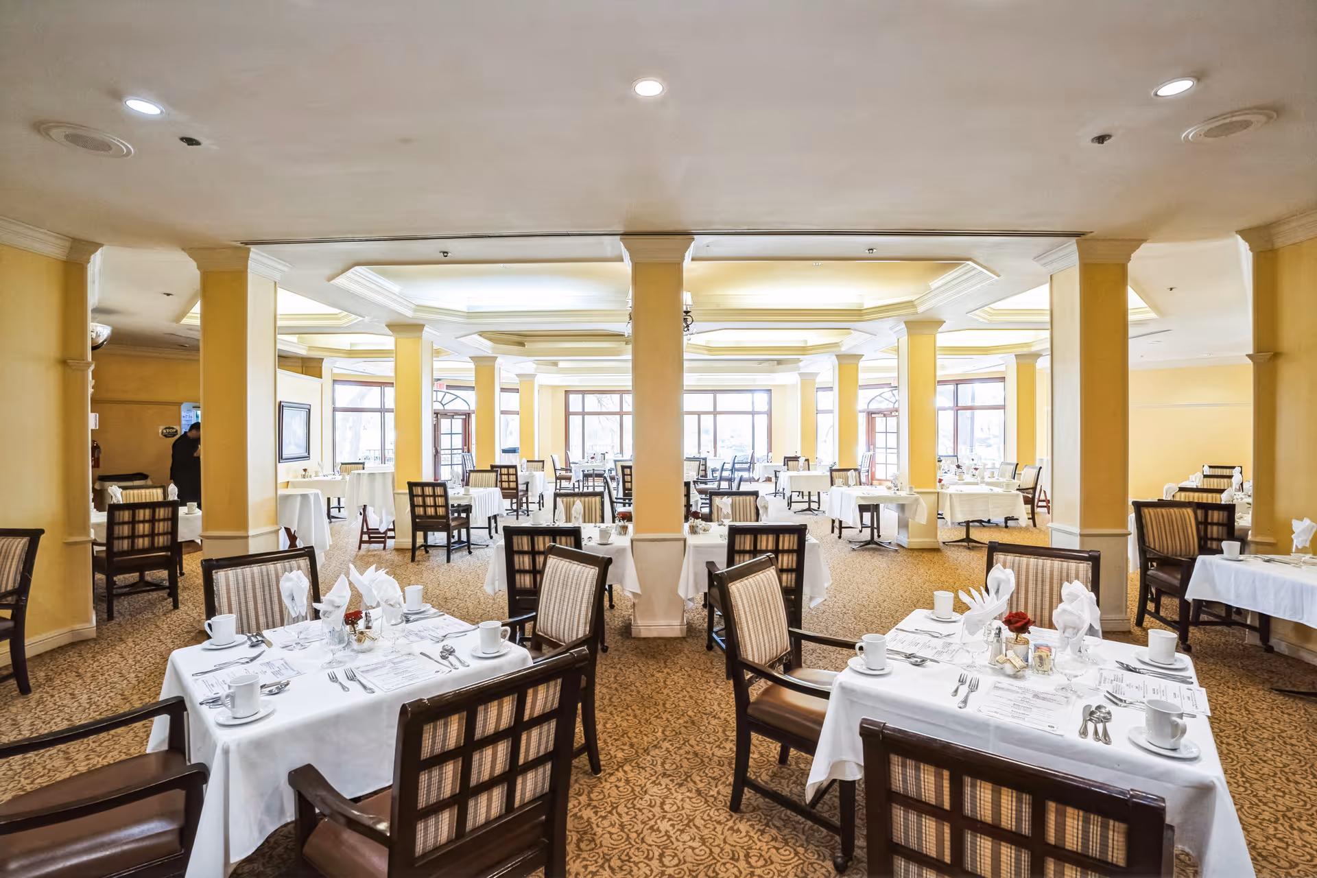Spacious dining room with multiple tables covered in white tablecloths, set with cups, napkins, and silverware. The room features yellow walls, large windows allowing natural light, and cushioned chairs with wooden frames arranged around the tables.