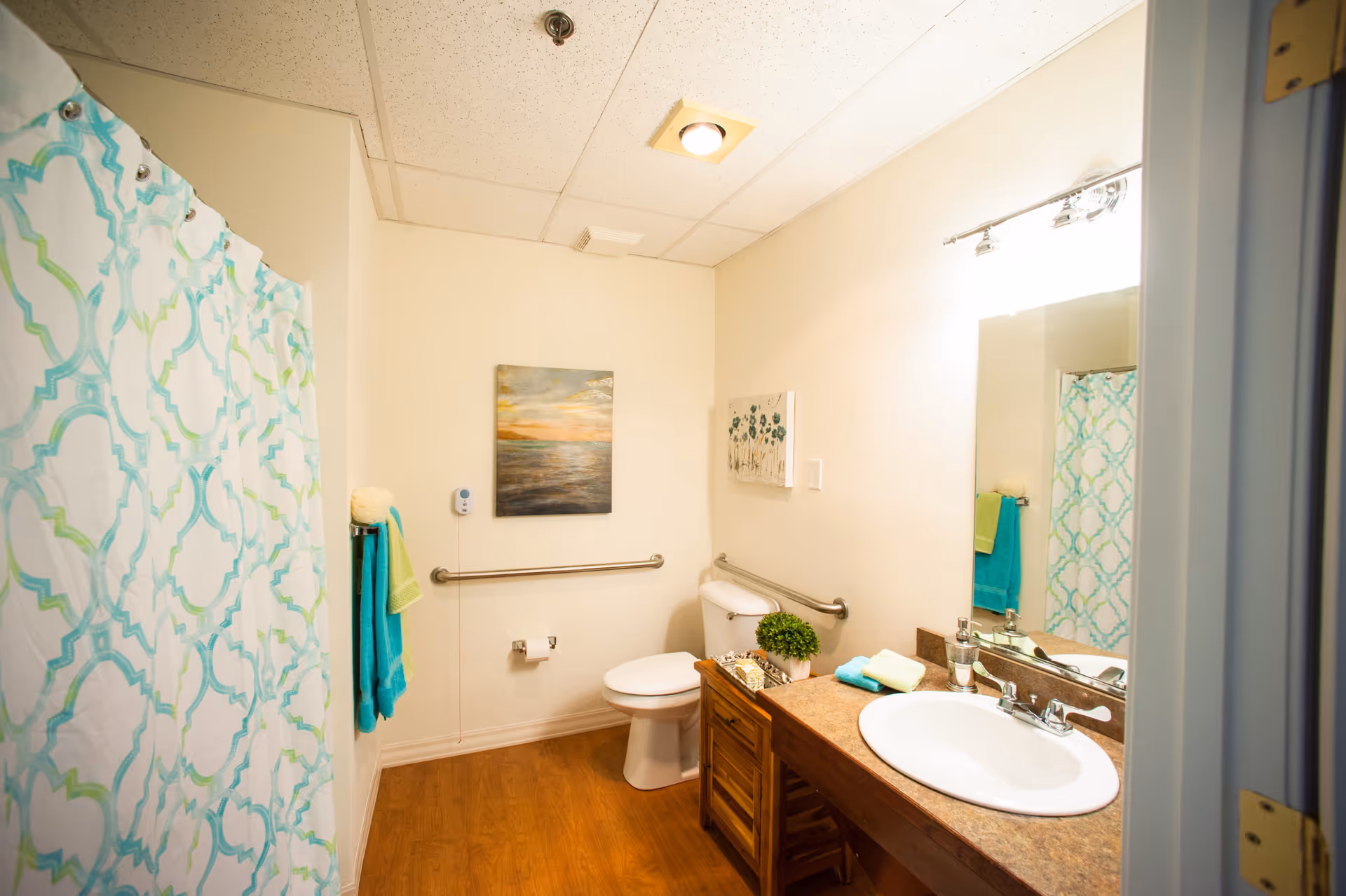 A clean and well-lit bathroom with a white toilet, a wooden vanity with a white sink, and a large mirror above it. The bathroom features a shower with a white and turquoise patterned curtain, wooden flooring, and light-colored walls decorated with two paintings. There are grab bars near the toilet and a towel rack with turquoise and green towels.