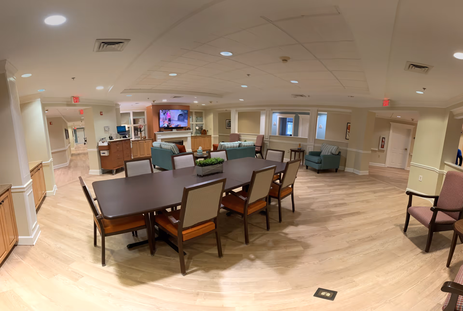 A spacious common area in a senior living facility featuring a large dark wood table with eight chairs in the foreground. In the background, there are two teal sofas and two armchairs arranged around a fireplace with a mounted television above it. The room has light wood flooring, beige walls with white trim, and recessed ceiling lights. Hallways and additional seating areas are visible in the distance.