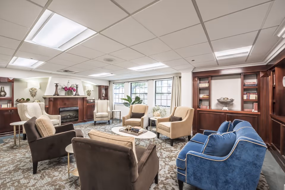A bright and spacious living room with a mix of beige, brown, and blue upholstered chairs and sofas arranged around a white circular coffee table. The room features a patterned carpet, large windows with beige curtains, built-in wooden bookshelves, and a fireplace with decorative items on the mantel.