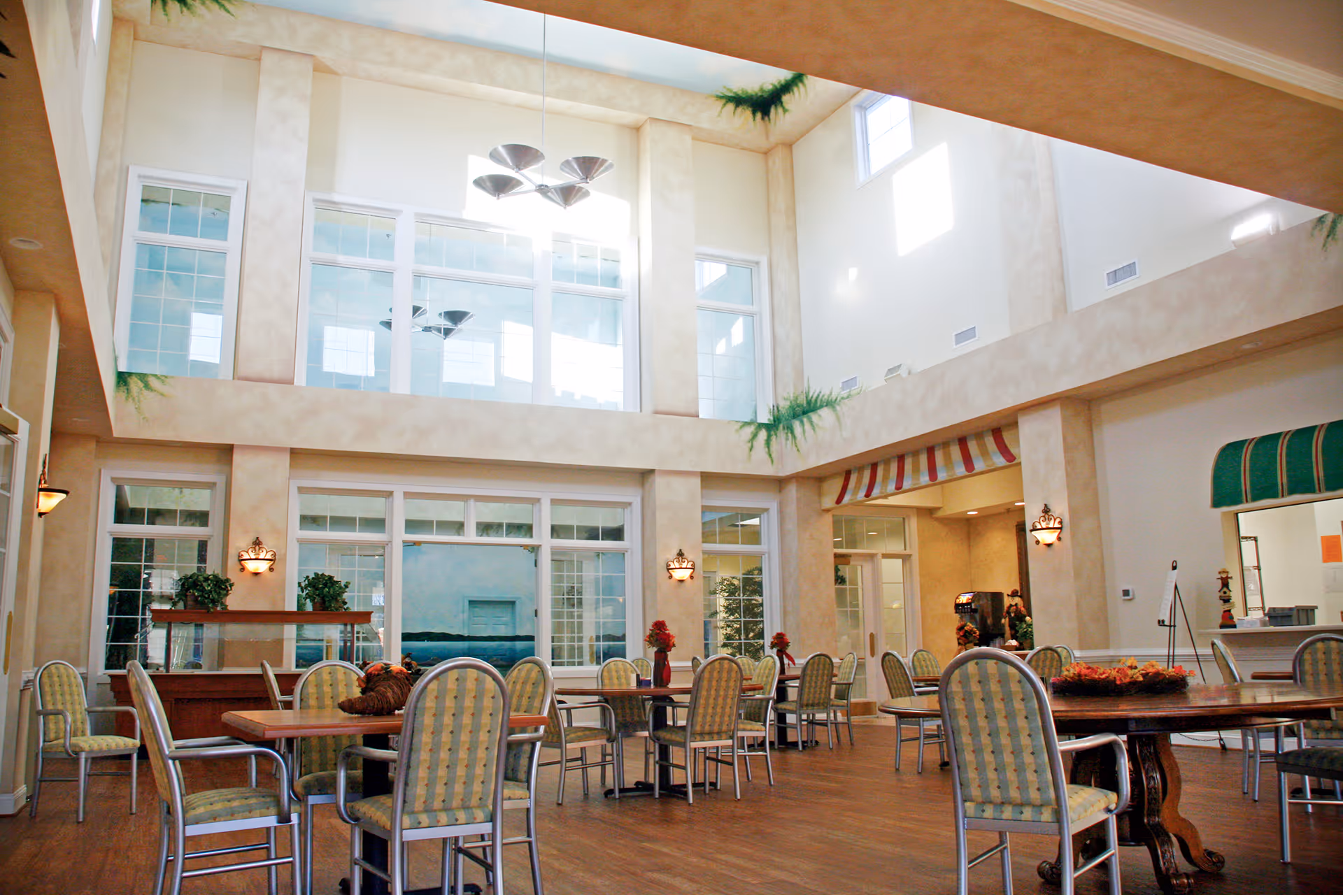 A spacious dining area with multiple tables and chairs arranged neatly. The room has high ceilings with large windows allowing natural light to fill the space. There are decorative plants and wall sconces, and the floor is wooden. The area appears clean and inviting.