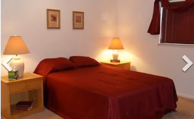 Simple bedroom with a double bed covered in dark red bedding, two bedside tables with lamps, framed wall art, and a curtained window.