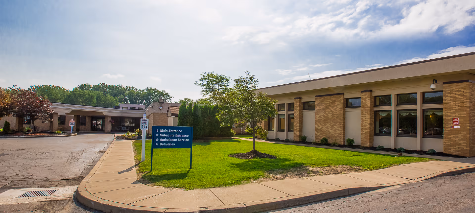 Front exterior of a single-story senior care facility with a driveway, lawn, tree, and a blue directional sign near the entrance.