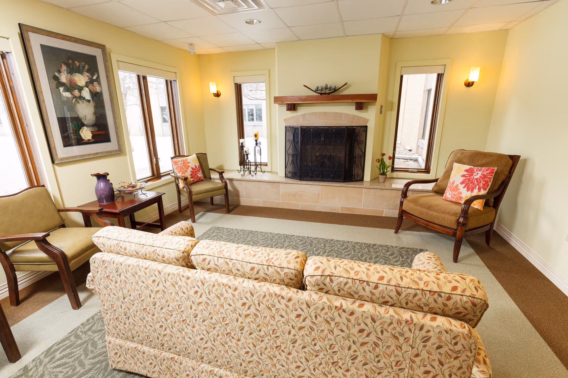 A cozy living room with a floral patterned sofa facing a fireplace with a decorative screen. The room has three armchairs, two with floral cushions, a side table with a purple vase and a bowl, and three tall windows letting in natural light. The walls are painted light yellow and there is a framed floral painting on one wall.