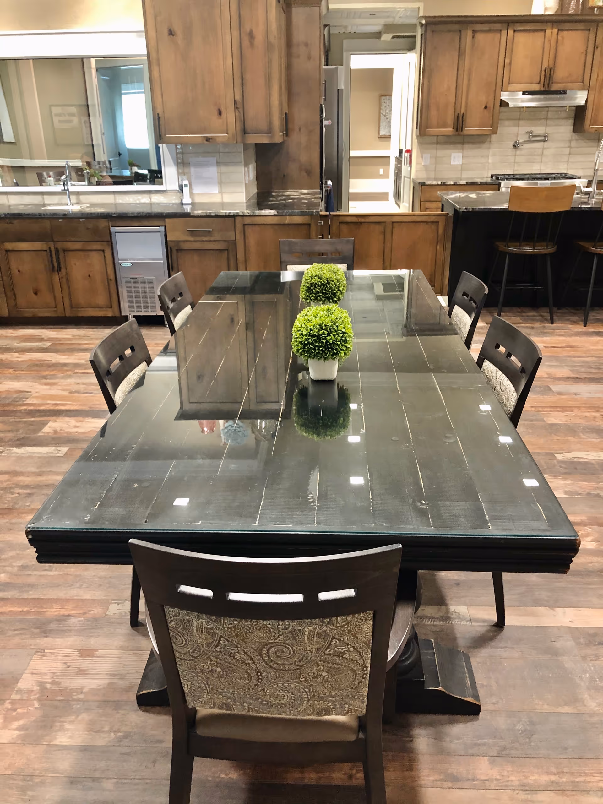 A dining area with a large rectangular glass-top table surrounded by eight wooden chairs with patterned cushions. Two small green potted plants are placed in the center of the table. In the background, there is a kitchen with wooden cabinets, a countertop, and bar stools.
