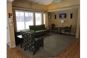A cozy living room area with a green sofa, a patterned armchair, a small table with two chairs, a wall-mounted TV, and large windows letting in natural light.