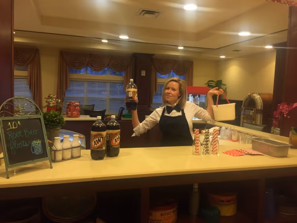 A woman in a black apron stands behind a counter holding a bottle of root beer and a small bucket at a beverage station with soda bottles and straws.