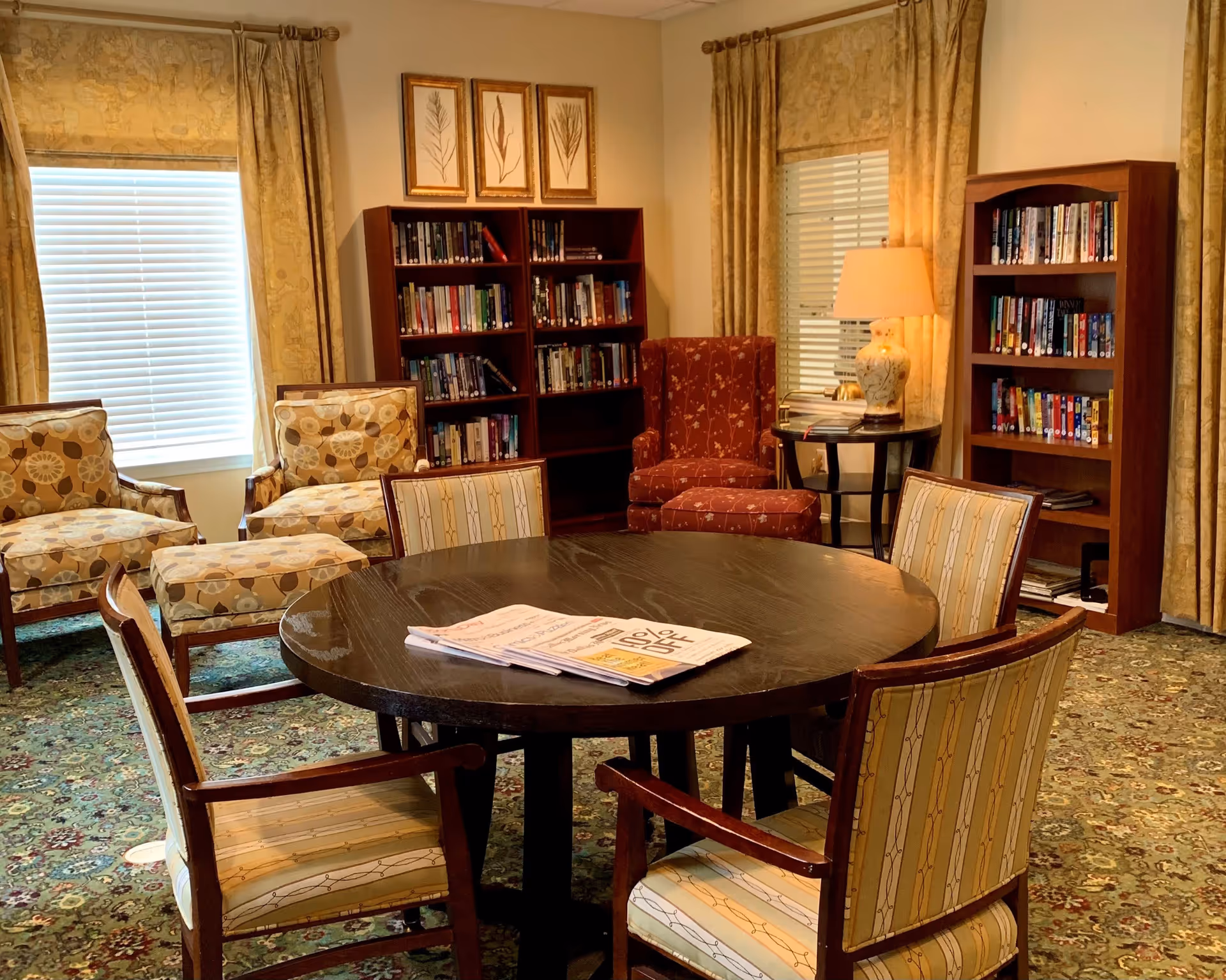 A cozy reading and sitting area in a senior living facility featuring a round wooden table with four upholstered chairs, two patterned armchairs with matching ottomans, a red armchair, two wooden bookshelves filled with books, a side table with a decorative lamp, and windows with beige curtains.