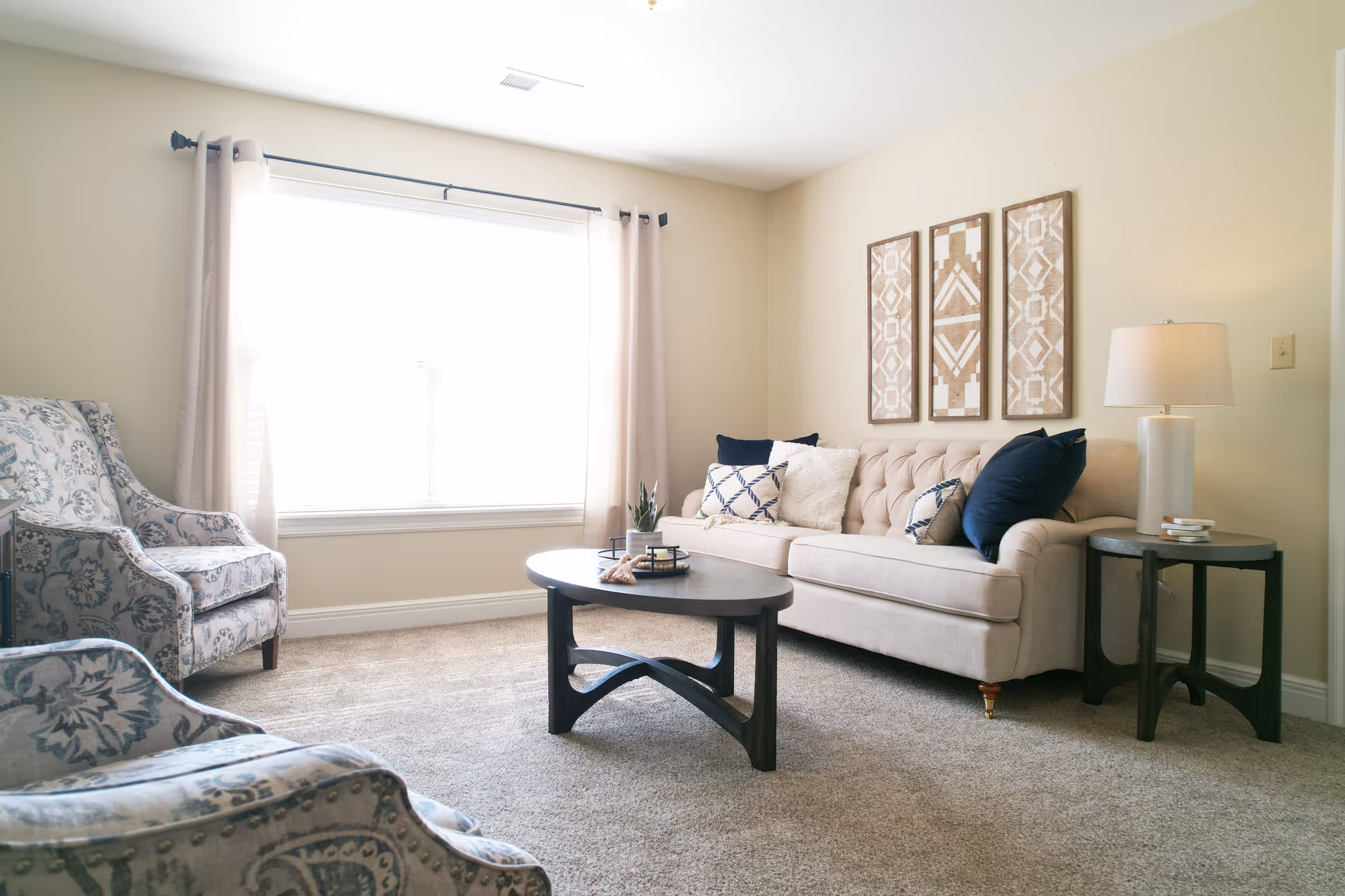 A bright living room with a large window covered by light curtains. The room features a beige tufted sofa with multiple cushions, two patterned armchairs, a dark wooden coffee table with decorative items, and a matching side table with a lamp. Three framed geometric art pieces hang on the wall above the sofa.