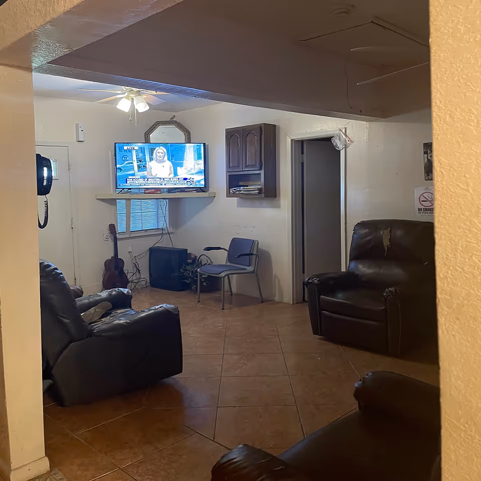 Small communal living room with leather recliners, a wall-mounted TV, a guitar, and a chair near a doorway.
