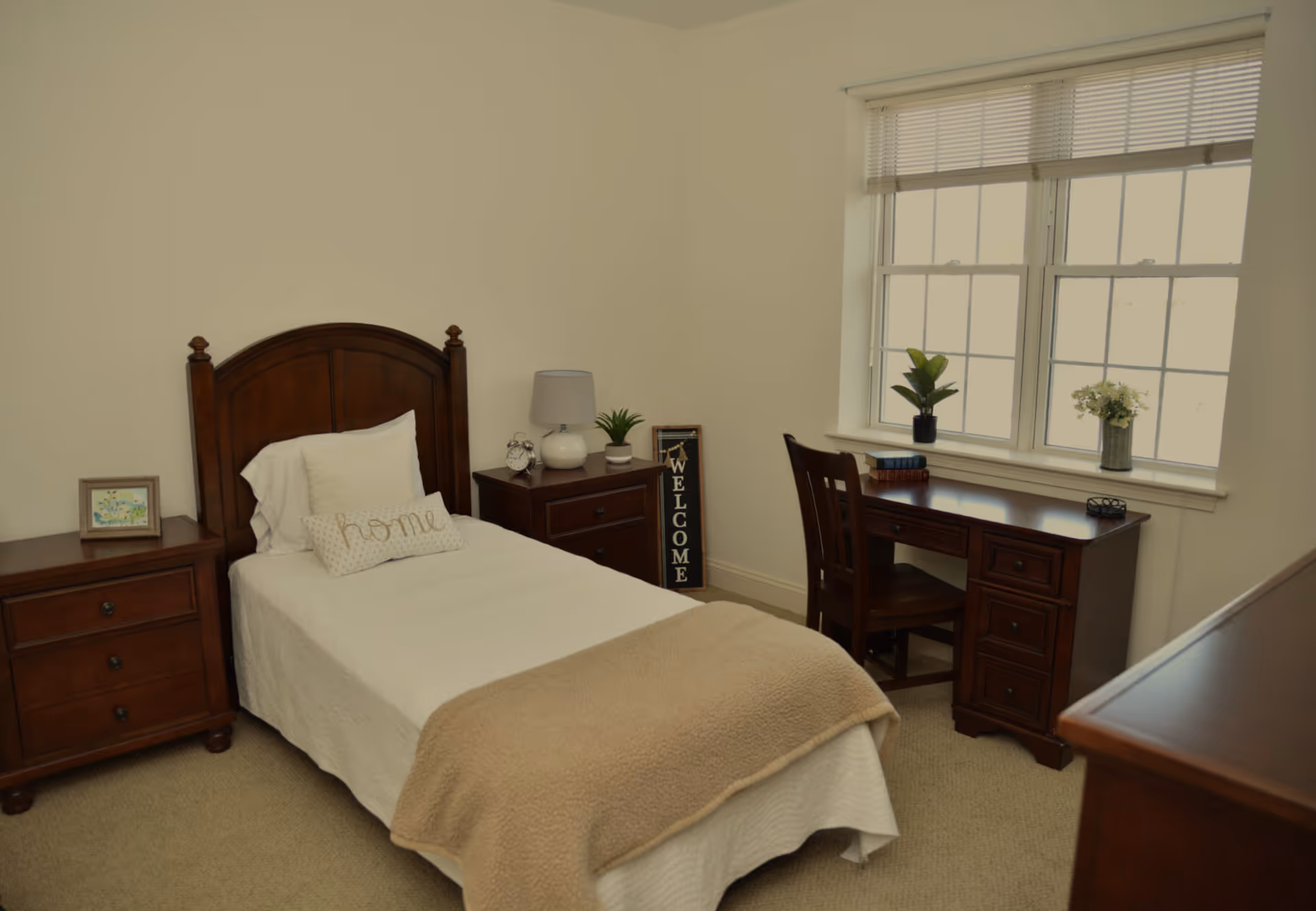 A neatly arranged bedroom with a single bed, wooden headboard and nightstands, a desk by a window, and neutral decor.