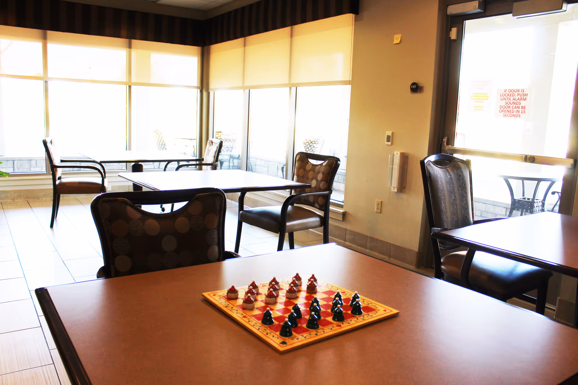 A well-lit room with several tables and chairs arranged around the space. On the closest table, there is a chess/checkers board with game pieces set up. Large windows with blinds allow natural light to fill the room, and a glass door leads outside to a patio area with additional seating.
