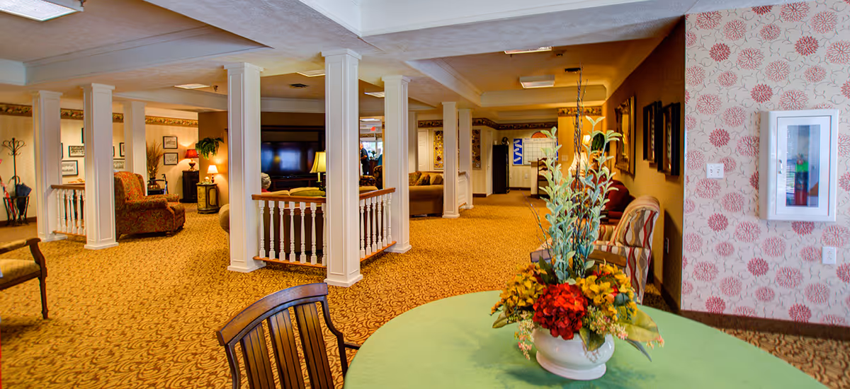 Interior view of a senior living facility common area with patterned carpet, white columns, and various seating arrangements including armchairs and sofas. A round table with a green tablecloth and a floral centerpiece is in the foreground. The walls have framed pictures and floral wallpaper on one side. The space is warmly lit with lamps and ceiling lights.
