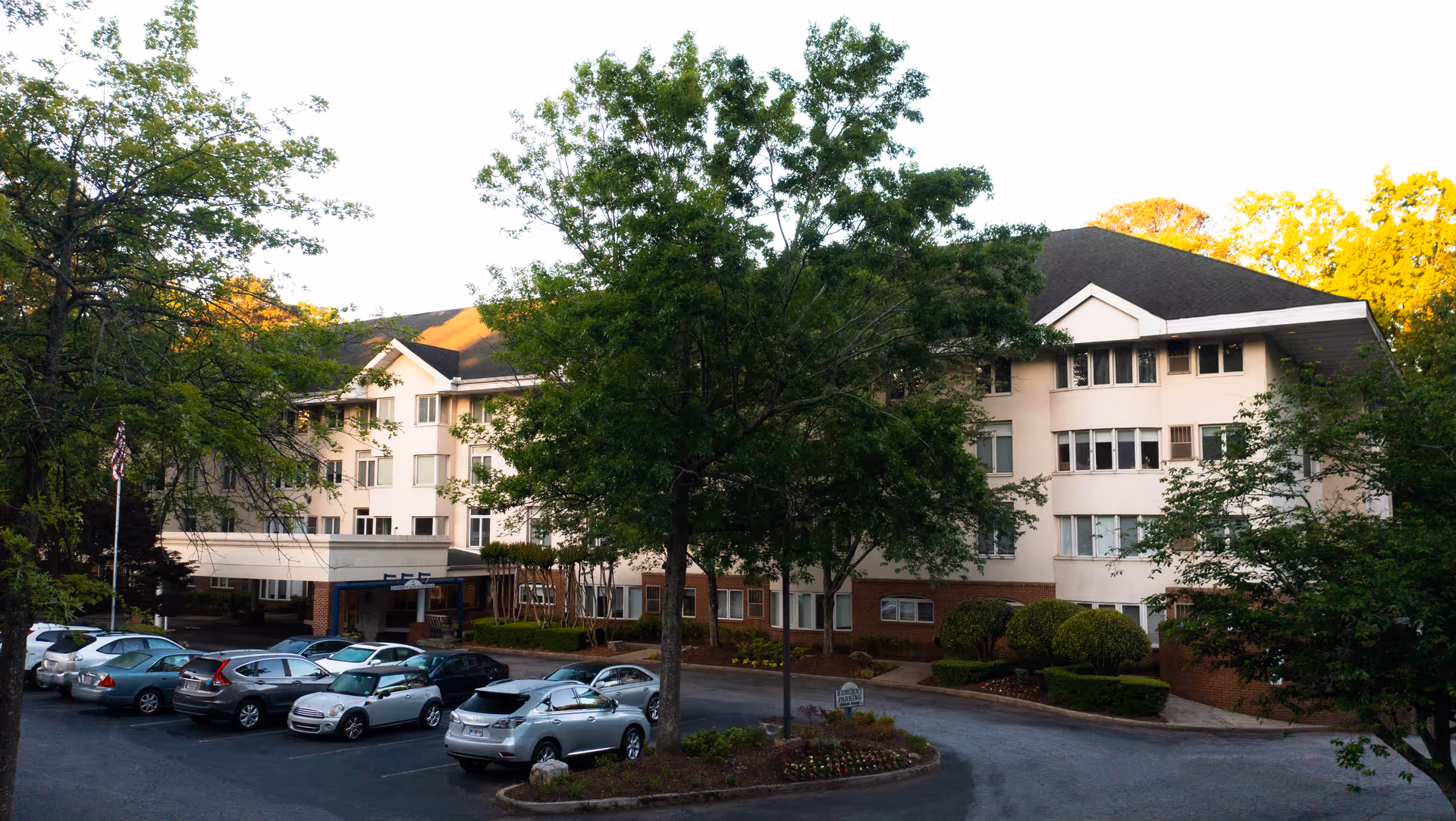 Exterior view of a multi-story senior living facility named Saint Anne's Terrace surrounded by trees and a parking lot with several cars parked.