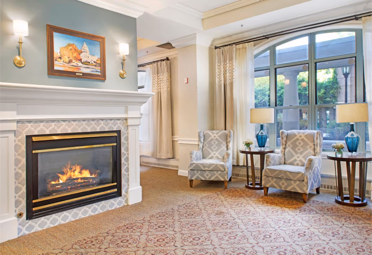 A cozy living room area featuring a lit fireplace with a decorative tile surround and white mantel. Above the fireplace is a framed painting of a capitol building with autumn trees. Two patterned armchairs are positioned near large arched windows with beige curtains. Each chair is accompanied by a small round wooden side table with a blue glass lamp and a small flower arrangement. The room has a patterned carpet and soft lighting from wall sconces and lamps.