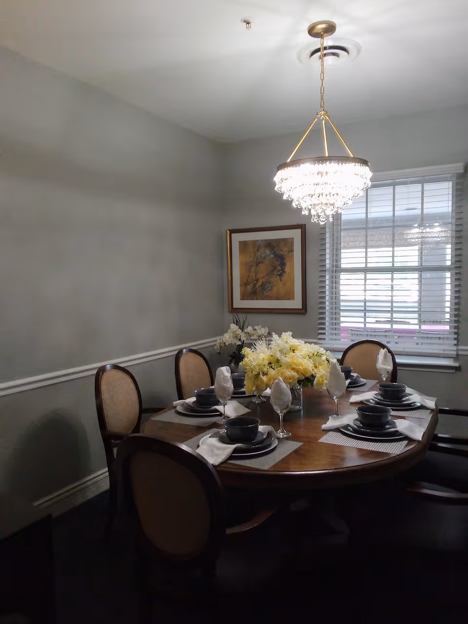 A dining room with a wooden oval table set for six people. Each place setting includes a dark bowl, plate, napkin, and a wine glass. A chandelier with crystal decorations hangs above the table. There is a window with blinds on the right wall and a framed painting on the back wall. A floral centerpiece with yellow and white flowers is on the table.
