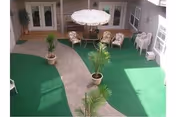 Outdoor courtyard area with green artificial turf, a curved concrete pathway, potted plants, a round table with an umbrella, and several chairs arranged around the table and along the walls.
