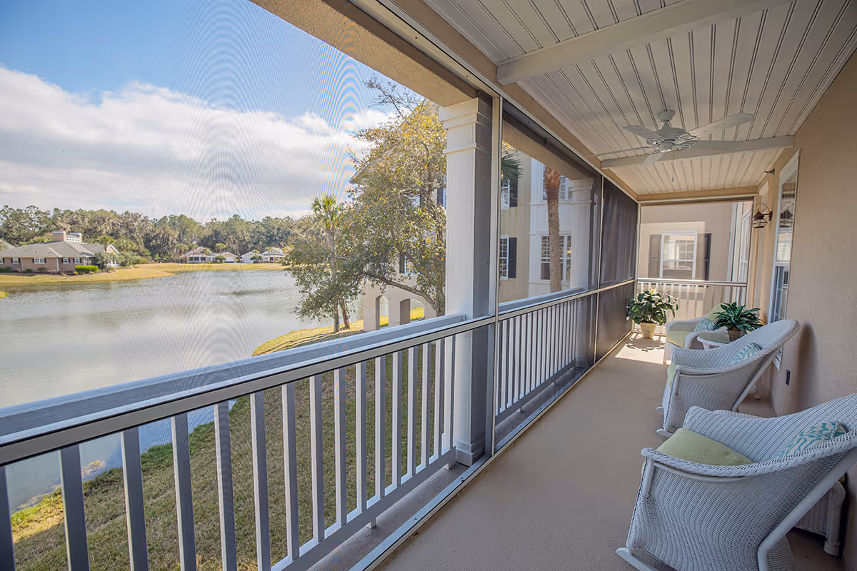 A screened-in balcony with white wicker chairs and potted plants overlooking a serene lake and houses in the distance under a partly cloudy sky.