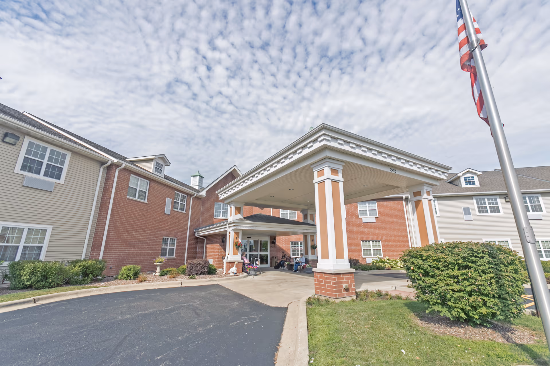 Exterior view of a senior living facility named Heritage Woods of Yorkville, showing a covered entrance with columns, a driveway, landscaped bushes, and an American flag on a flagpole under a partly cloudy sky.