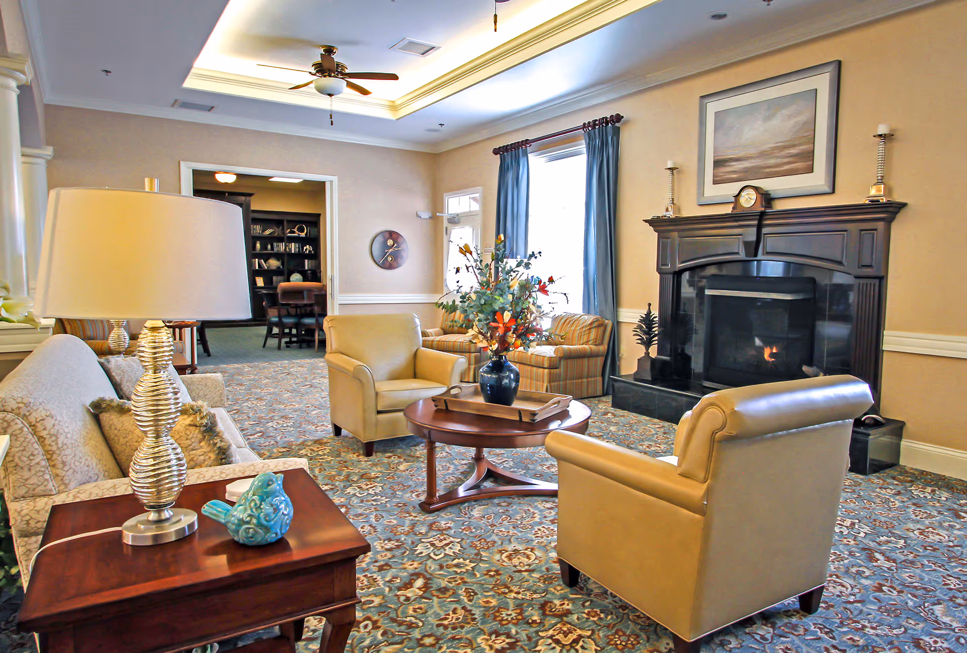 Bright, well-appointed common living room with upholstered chairs, a sofa, coffee table with a floral arrangement, and a lit fireplace.