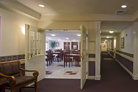 View of an interior hallway leading to a dining area with tables and chairs. The hallway has carpeted floors and framed artwork on the walls. Double doors are open, revealing the dining room with a tiled floor and more seating. The space is well-lit with ceiling lights.
