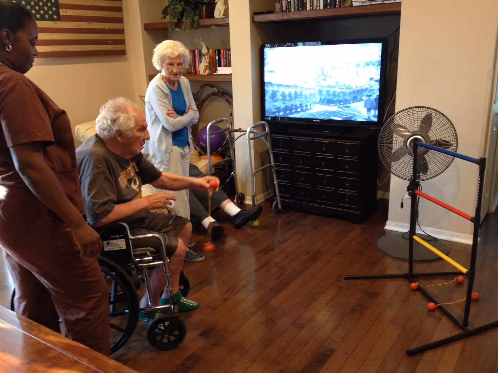 An elderly man in a wheelchair playing a game with a ball and a ladder-like structure, assisted by a caregiver in brown scrubs. An elderly woman stands nearby watching and smiling. The room has wooden floors, a TV on a black cabinet, a fan, and an American flag on the wall.
