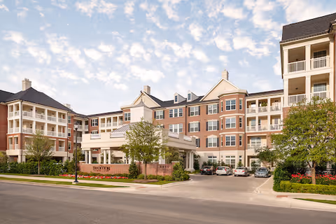 Exterior view of The Tradition-Lovers Lane senior living facility showing a large multi-story brick building with balconies, a covered entrance, landscaped greenery, and parked cars in front under a partly cloudy sky.
