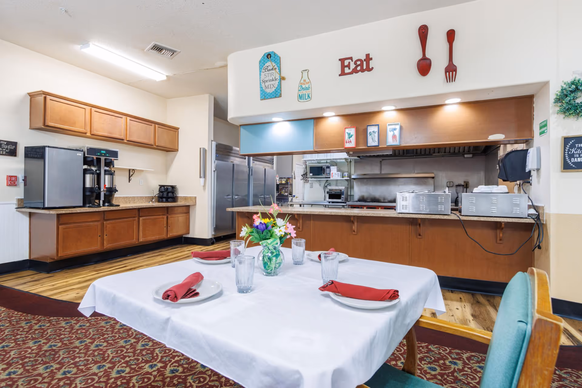 Table set with plates, glasses and napkins in a dining area facing a serving counter and kitchen service window.