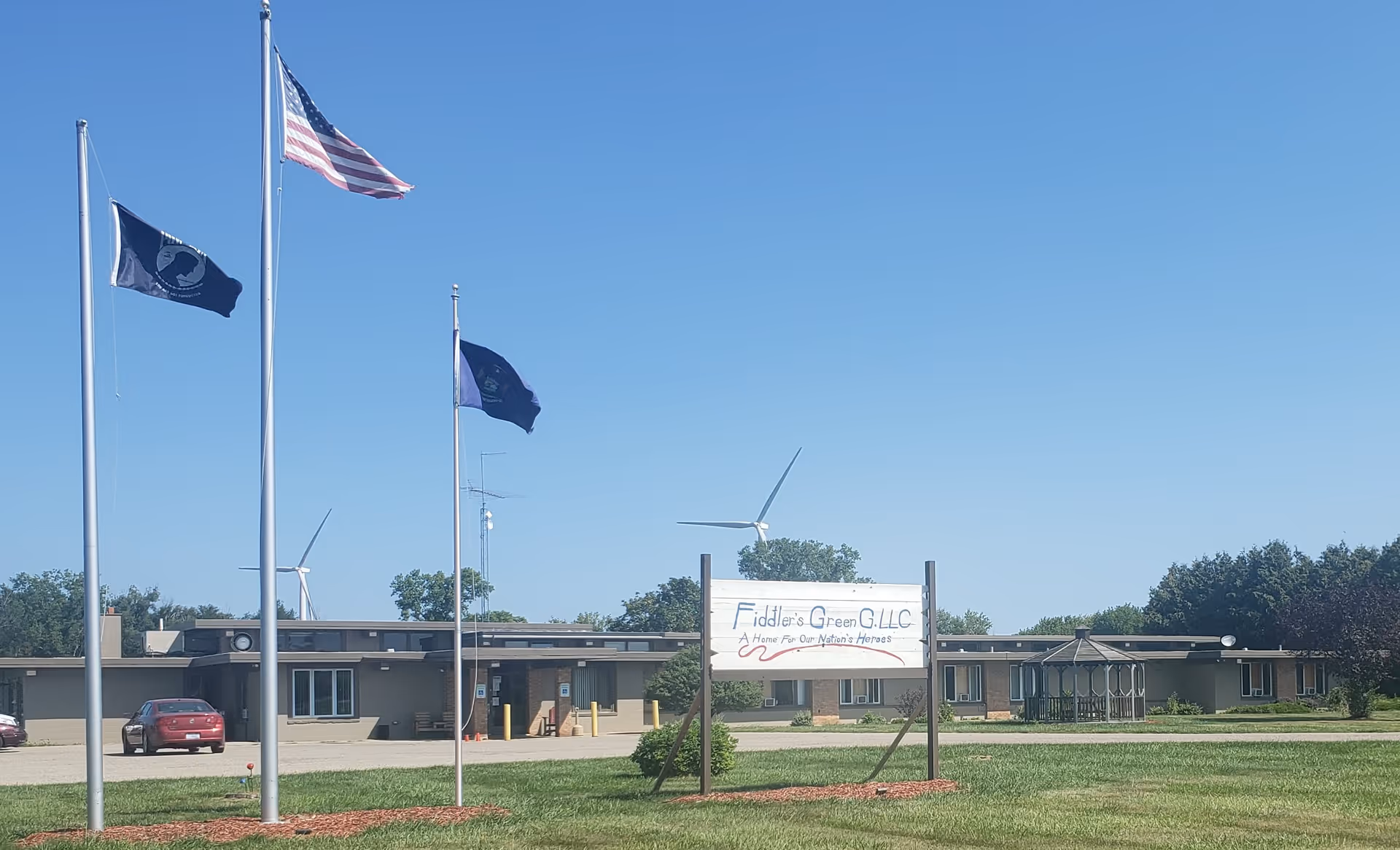 Exterior view of Fiddler's Green G. LLC senior community building with three flagpoles flying flags, a sign in front, a gazebo to the right, and a clear blue sky with wind turbines in the background.