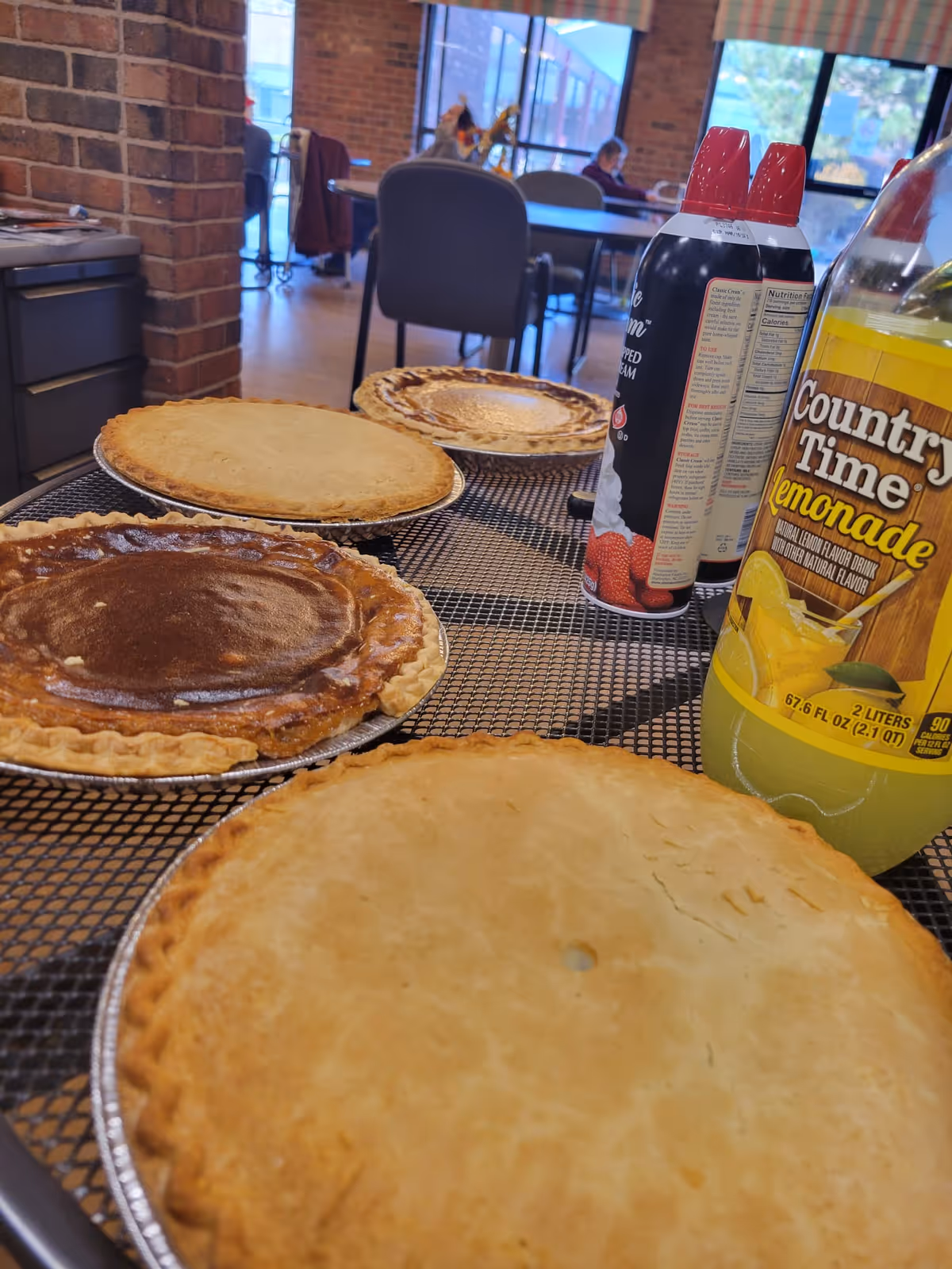 Four pies placed on a metal mesh table with two cans of whipped cream and a bottle of Country Time lemonade. In the background, there are chairs and tables in a room with large windows and brick walls.