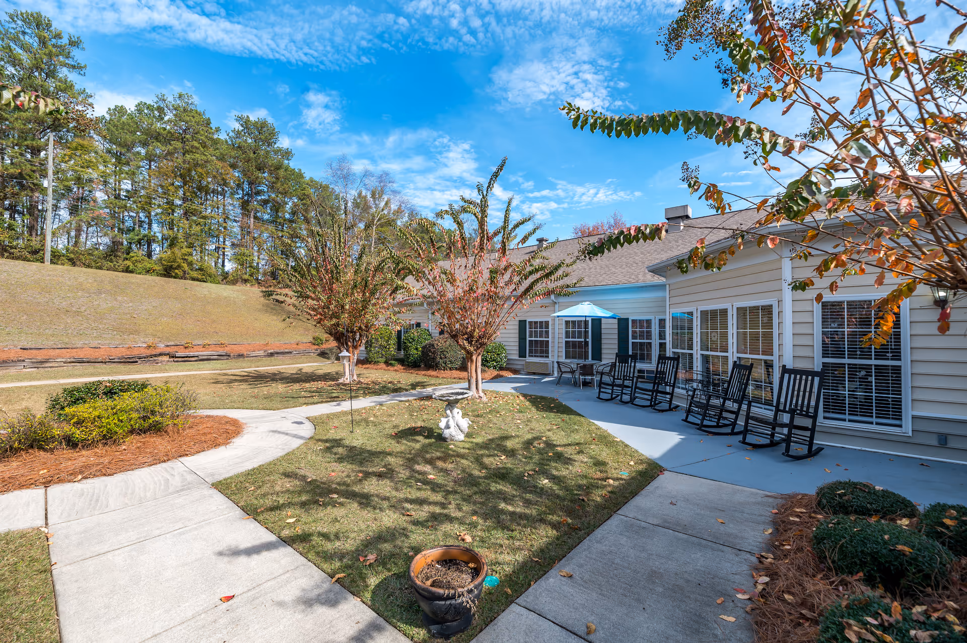 Outdoor patio area at Oaks at Maple Ridge featuring a concrete walkway, several black rocking chairs, a table with a blue umbrella, small trees, shrubs, and a clear blue sky with some clouds.