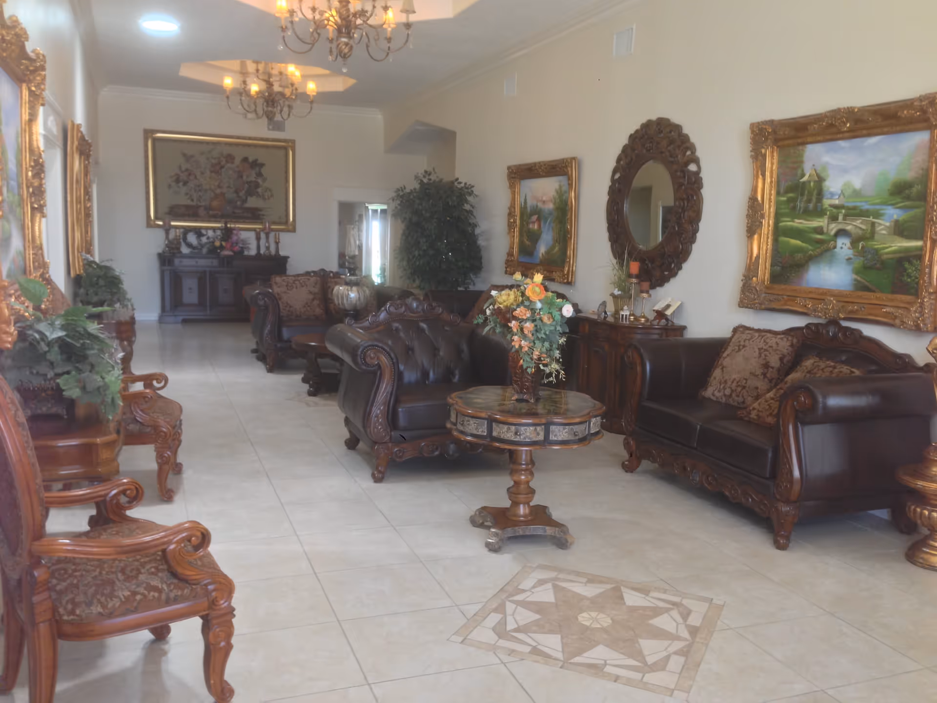 A spacious and elegantly decorated living room with classic dark leather sofas, ornate wooden chairs, and a round wooden coffee table with a floral arrangement. The room features tiled floors with a decorative tile pattern, framed paintings, a large round mirror on the wall, chandeliers hanging from the ceiling, and various plants and decorative items.