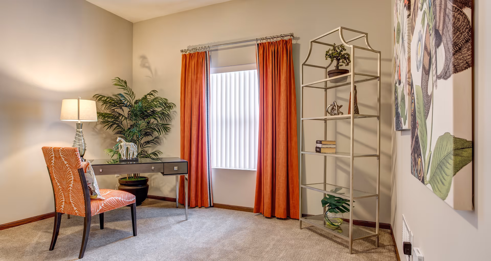 A cozy interior room with beige walls and carpeted floor featuring a small black desk with a decorative horse figurine, an orange patterned chair, a tall potted plant, and a floor lamp. There is a window with vertical blinds and orange curtains. On the right side, there is a metal shelving unit with decorative items and a large floral artwork on the wall.